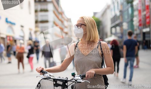 Image of Woman walking by her bicycle on pedestrian city street wearing medical face mask in public to prevent spreading of corona virus. New normal during covid epidemic.