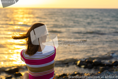 Image of A girl sits in the evening at sunset on the seashore, view from the back