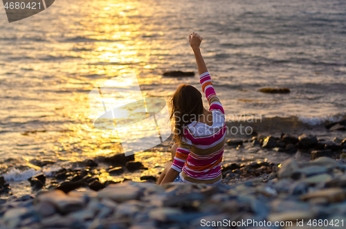 Image of A girl sits on the seashore and throws stones into the water, raised her hands, evening, sunset