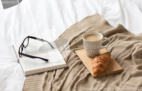 Image of coffee, croissant, blanket and book on bed at home