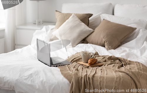 Image of laptop, cup of coffee and croissant on bed at home