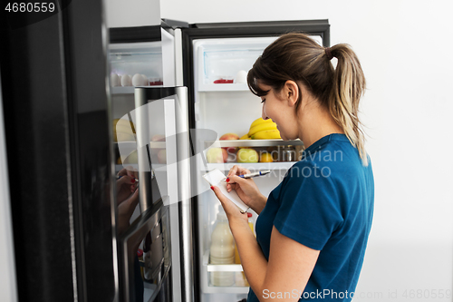 Image of woman making list of necessary food at home fridge