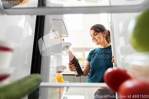 Image of happy woman taking wine bottle from fridge at home