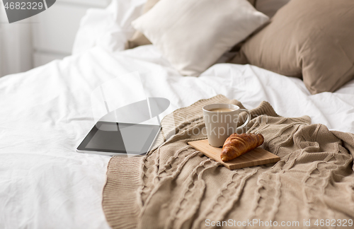 Image of tablet pc, coffee cup and croissant on bed at home