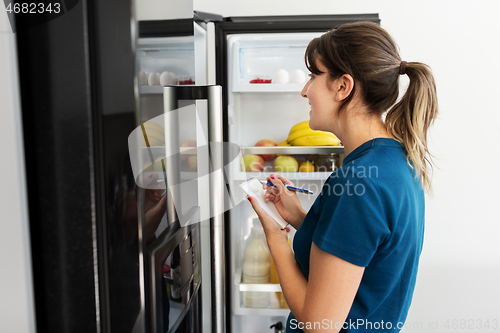 Image of woman making list of necessary food at home fridge