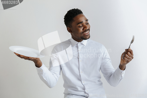 Image of Young smiling attractive african american guy holding empty dish and fork isolated on grey background. Copy space and mock up. Blank template background.