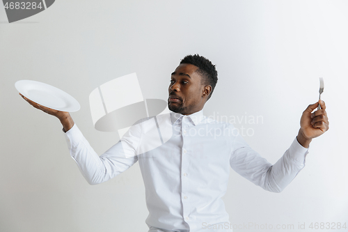 Image of Young doubting attractive african american guy holding empty dish and fork isolated on grey background. Copy space and mock up. Blank template background.