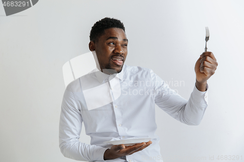 Image of Young african american guy holding empty dish and fork with disgusting facial expression isolated on grey background. Copy space and mock up. Blank template background.