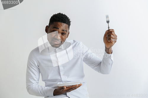 Image of Young doubting attractive african american guy holding empty dish and fork isolated on grey background. Copy space and mock up. Blank template background.