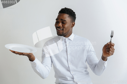 Image of Young doubting attractive african american guy holding empty dish and fork isolated on grey background. Copy space and mock up. Blank template background.