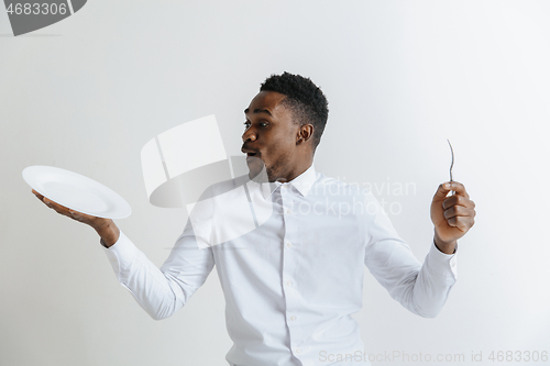 Image of Young surprised attractive african american guy holding empty dish and fork isolated on grey background. Copy space and mock up. Blank template background.