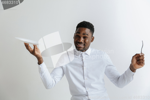 Image of Young doubting attractive african american guy holding empty dish and fork isolated on grey background. Copy space and mock up. Blank template background.