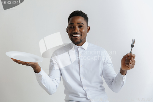 Image of Young smiling attractive african american guy holding empty dish and fork isolated on grey background. Copy space and mock up. Blank template background.