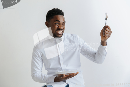 Image of Young african american guy holding empty dish and fork with disgusting facial expression isolated on grey background. Copy space and mock up. Blank template background.