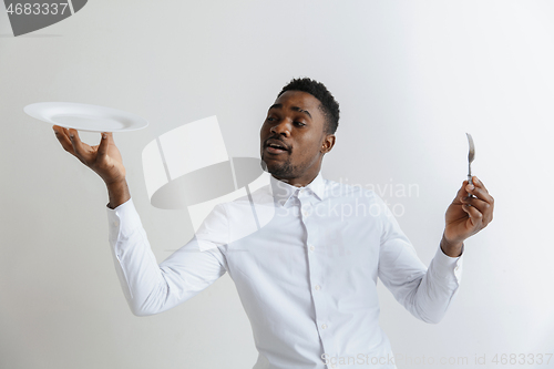 Image of Young doubting attractive african american guy holding empty dish and fork isolated on grey background. Copy space and mock up. Blank template background.