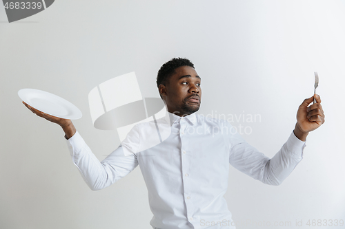 Image of Young doubting attractive african american guy holding empty dish and fork isolated on grey background. Copy space and mock up. Blank template background.