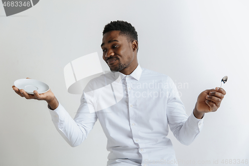 Image of Young doubting attractive african american guy holding empty dish and spoon isolated on grey background. Copy space and mock up. Blank template background.