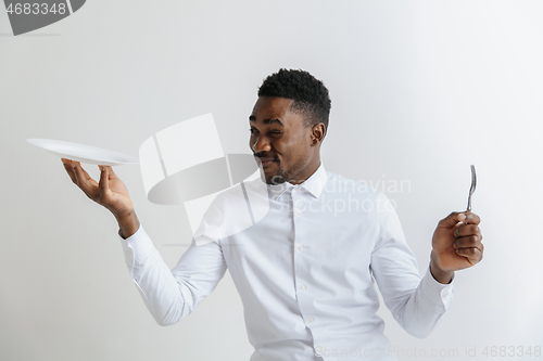 Image of Young doubting attractive african american guy holding empty dish and fork isolated on grey background. Copy space and mock up. Blank template background.