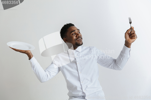 Image of Young doubting attractive african american guy holding empty dish and fork isolated on grey background. Copy space and mock up. Blank template background.