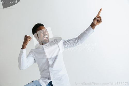 Image of Handsome Afro American man in classic shirt is smiling, looking at camera and pointing away, against white brick wall