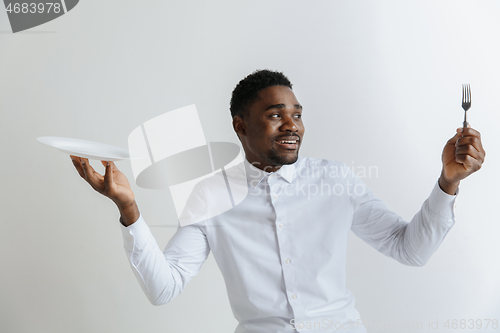 Image of Young doubting attractive african american guy holding empty dish and fork isolated on grey background. Copy space and mock up. Blank template background.