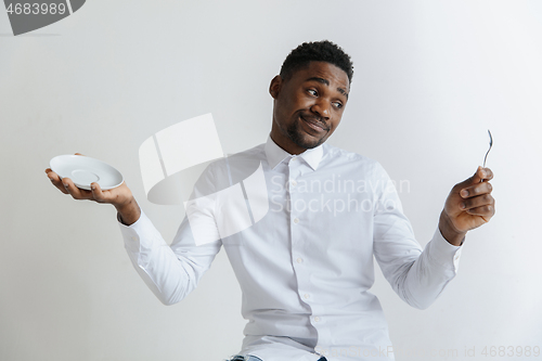 Image of Young doubting attractive african american guy holding empty dish and spoon isolated on grey background. Copy space and mock up. Blank template background.