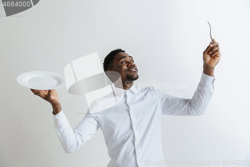 Image of Young doubting attractive african american guy holding empty dish and fork isolated on grey background. Copy space and mock up. Blank template background.