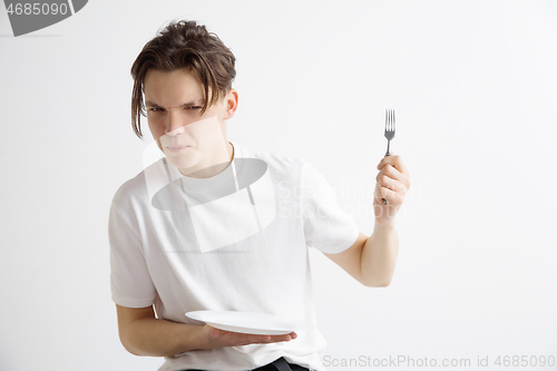 Image of Young sad attractive guy holding empty dish and fork isolated on grey background.