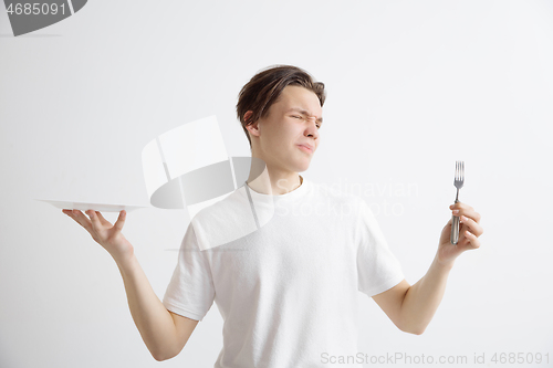 Image of Young sad attractive guy holding empty dish and fork isolated on grey background.