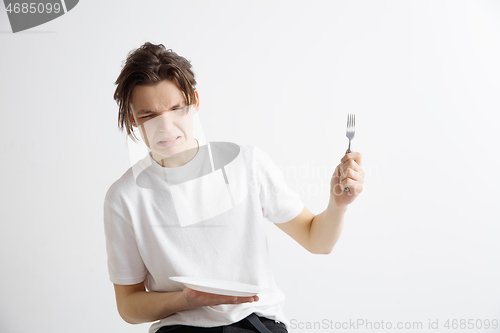 Image of Young sad attractive guy holding empty dish and fork isolated on grey background.