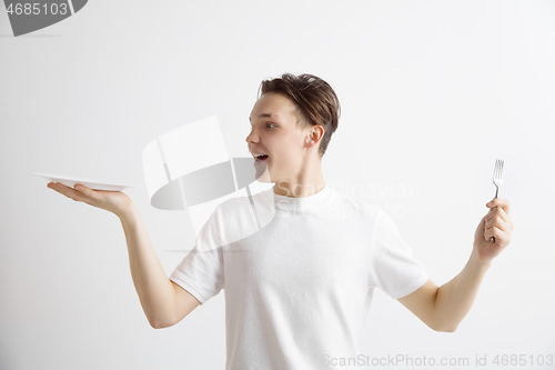 Image of Young smiling attractive guy holding empty dish and fork isolated on grey background.