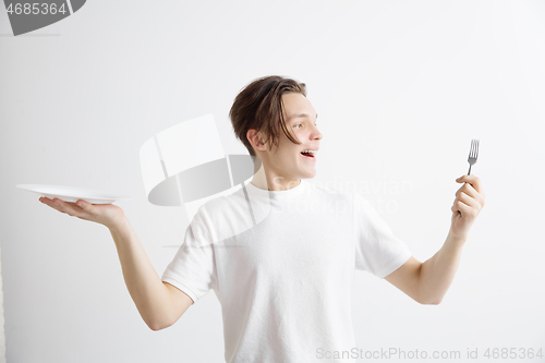 Image of Young smiling attractive guy holding empty dish and fork isolated on grey background.