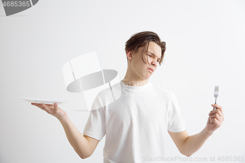 Image of Young sad attractive guy holding empty dish and fork isolated on grey background.