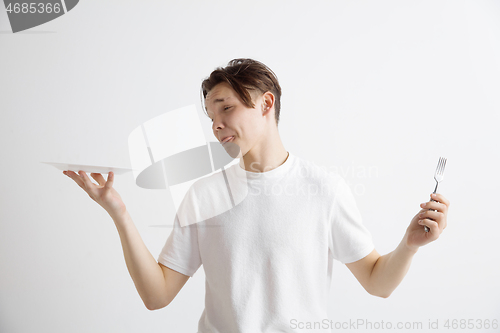 Image of Young sad attractive guy holding empty dish and fork isolated on grey background.