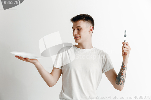 Image of Young smiling attractive guy holding empty dish and fork isolated on grey background.