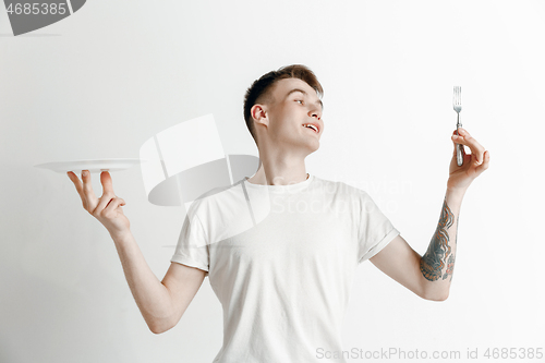 Image of Young smiling attractive guy holding empty dish and fork isolated on grey background.