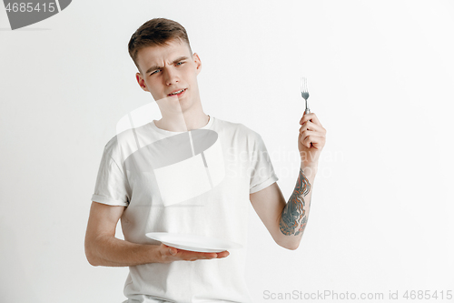 Image of Young sad attractive guy holding empty dish and fork isolated on grey background.