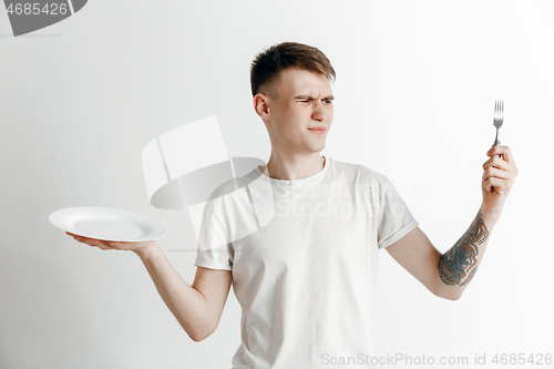 Image of Young sad attractive guy holding empty dish and fork isolated on grey background.