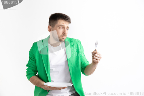 Image of Young sad attractive guy holding empty dish and fork isolated on grey background.