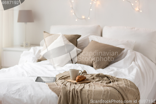 Image of tablet pc, coffee cup and croissant on bed at home