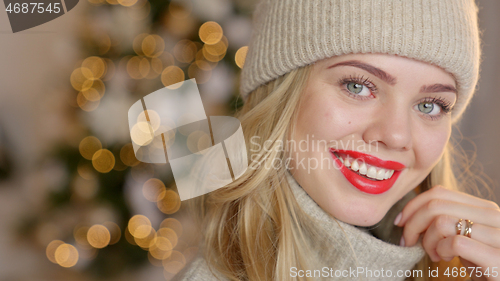 Image of Smiling blond woman with red lipstick, dressed in a warm woolen cardigan and hat posing in her home