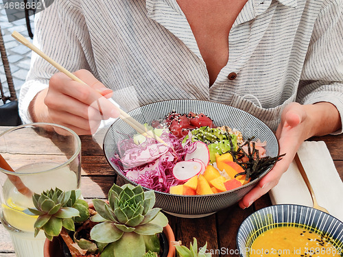 Image of Woman eating tasty colorful healthy natural organic vegetarian Hawaiian poke bowl using asian chopsticks on rustic wooden table. Healthy natural organic eating concept