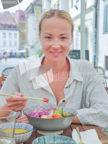 Image of Woman eating tasty colorful healthy natural organic vegetarian Hawaiian poke bowl using asian chopsticks on rustic wooden table. Healthy natural organic eating concept