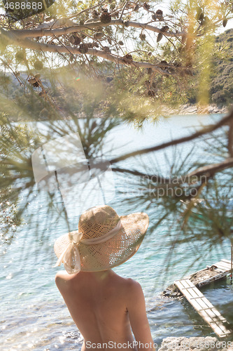 Image of Rear view of topless beautiful woman wearing nothing but straw sun hat realaxing on wild coast of Adriatic sea on a beach in shade of pine tree.