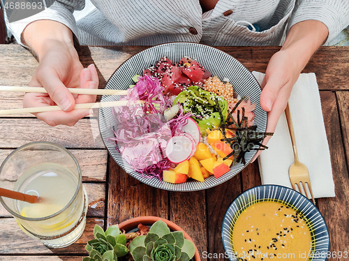 Image of Woman eating tasty colorful healthy natural organic vegetarian Hawaiian poke bowl using asian chopsticks on rustic wooden table. Healthy natural organic eating concept