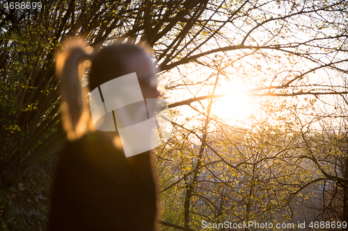 Image of Backlit defocused rear view of young woman talking on cell phone outdoors in park at sunset. Girl holding mobile phone, using digital device, looking at setting sun