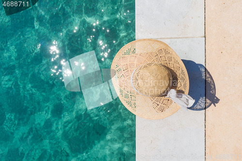 Image of Woman wearing big summer sun hat relaxing on pier by clear turquoise sea.