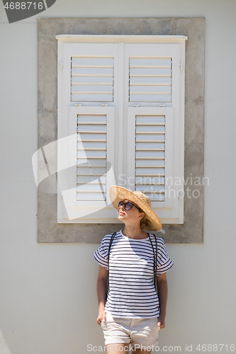 Image of Beautiful young female tourist woman wearing sun hat, standing and relaxing in front of vinatage wooden window in old Mediterranean town while sightseeing on hot summer day