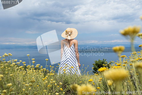 Image of Rear view of young woman wearing striped summer dress and straw hat standing in super bloom of wildflowers, relaxing while enjoing beautiful view of Adriatic sea nature, Croatia