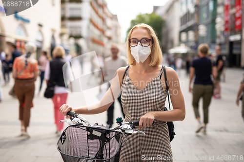 Image of Woman walking by her bicycle on pedestrian city street wearing medical face mask in public to prevent spreading of corona virus. New normal during covid epidemic.